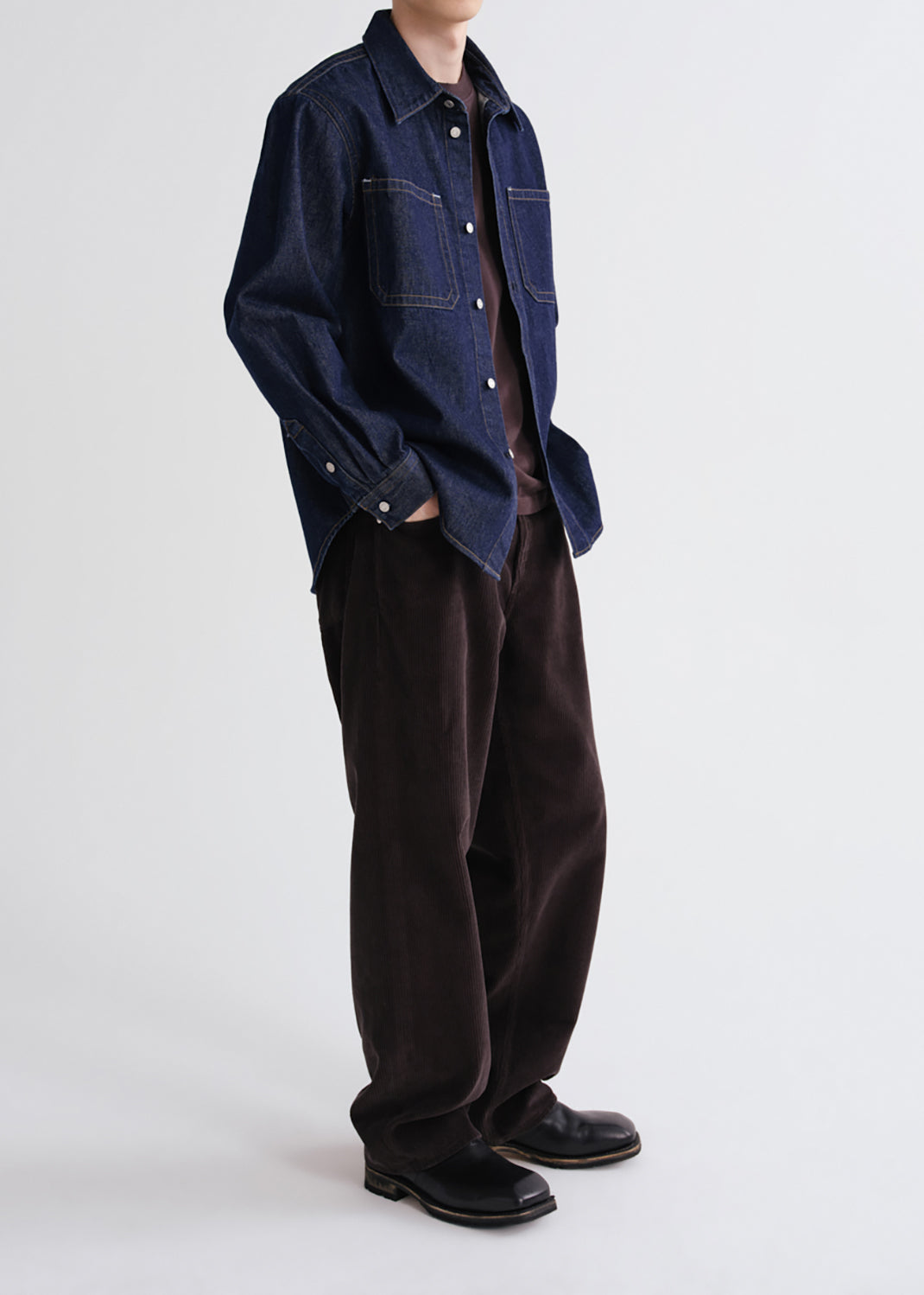 A model wearing a dark blue denim button-up shirt over a brown t-shirt, paired with dark brown corduroy trousers and black leather shoes, photographed against a grey background.