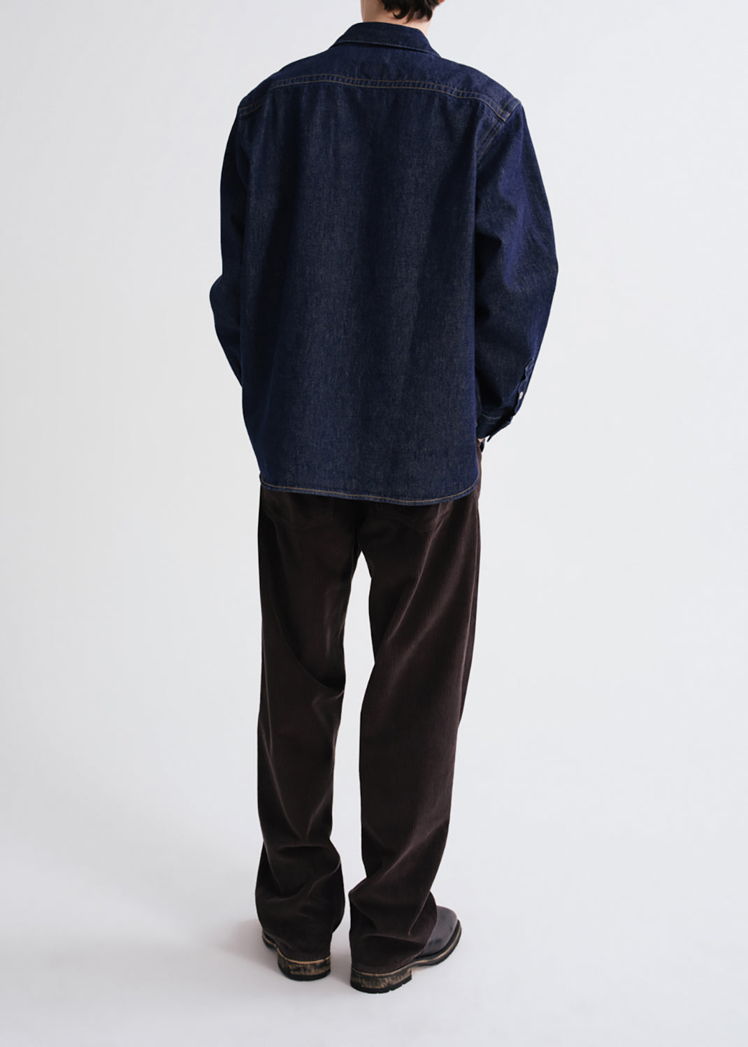 A person shown from the back wearing an oversized dark indigo denim shirt jacket and dark brown corduroy trousers, standing against a grey background.