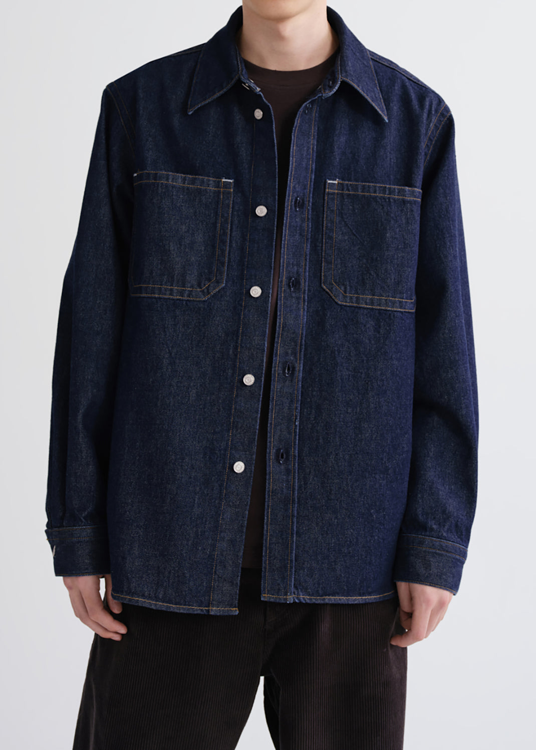 A person wearing a dark indigo denim button-up shirt jacket over a black t-shirt and black pants, shown from the neck down against a grey background.