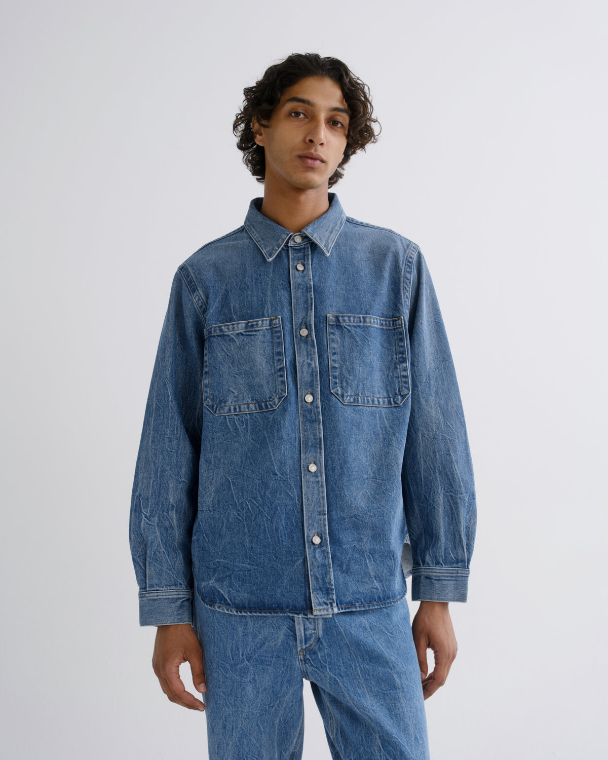 A man wearing a medium-wash blue denim button-up shirt and matching denim pants, standing against a plain light gray studio background.