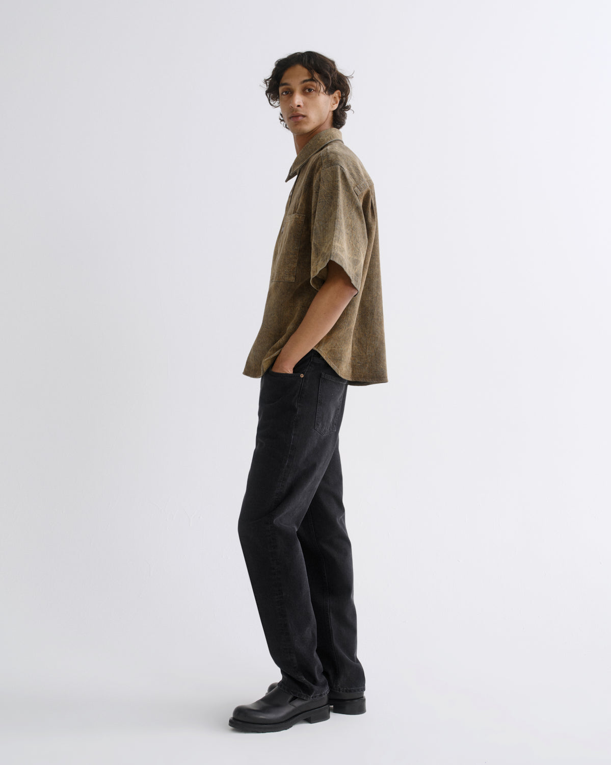 A model wearing a short-sleeve brown corduroy shirt and black jeans, standing in profile against a plain light gray studio background.