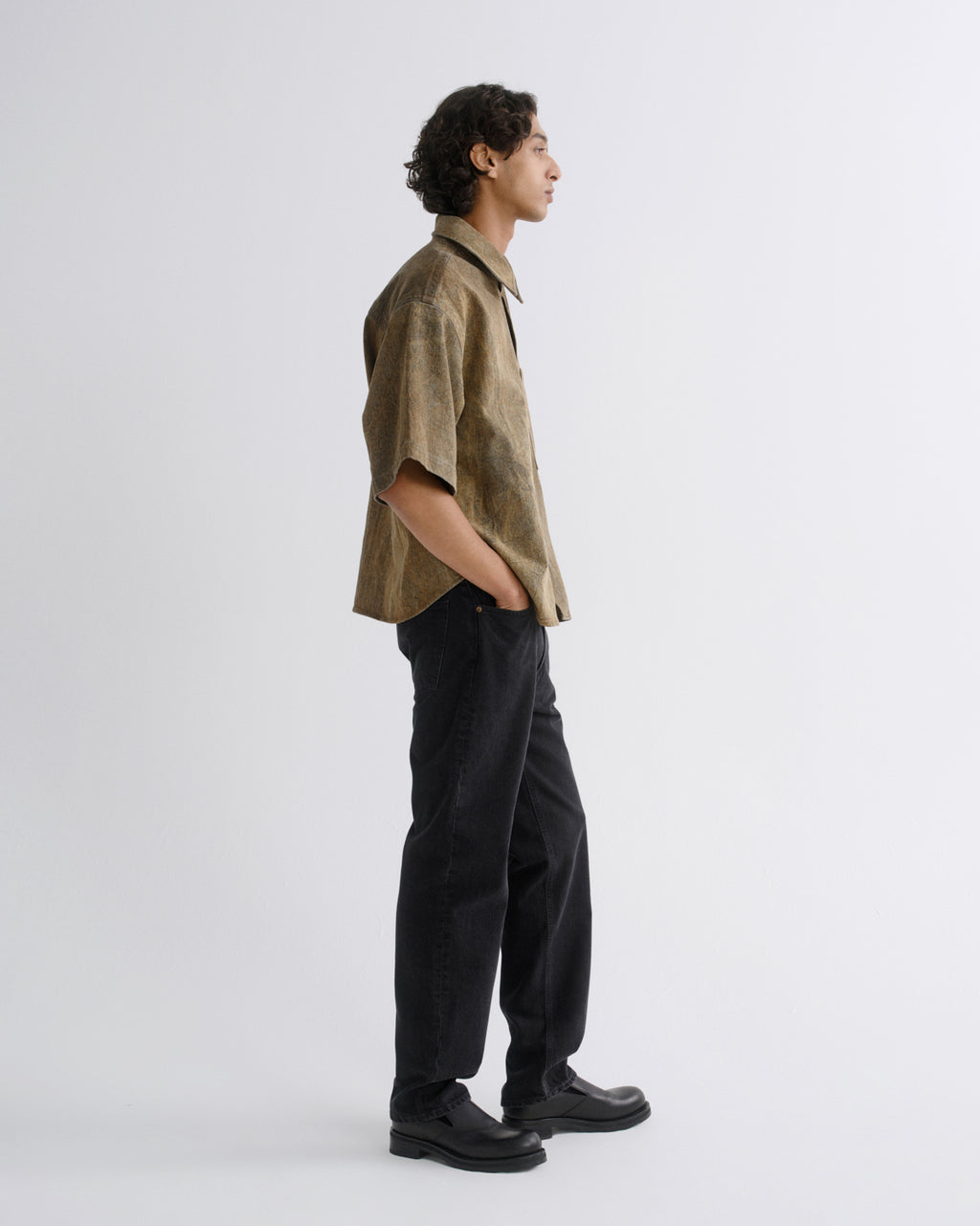 A man in a side profile wearing a short-sleeve button-up shirt in a textured khaki-brown fabric, paired with black trousers and black shoes, standing against a plain light gray studio background.
