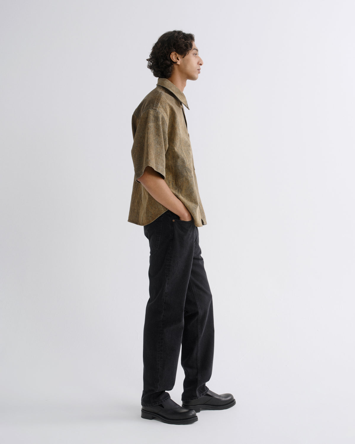 A man in a side profile wearing a short-sleeve button-up shirt in a textured khaki-brown fabric, paired with black trousers and black shoes, standing against a plain light gray studio background.