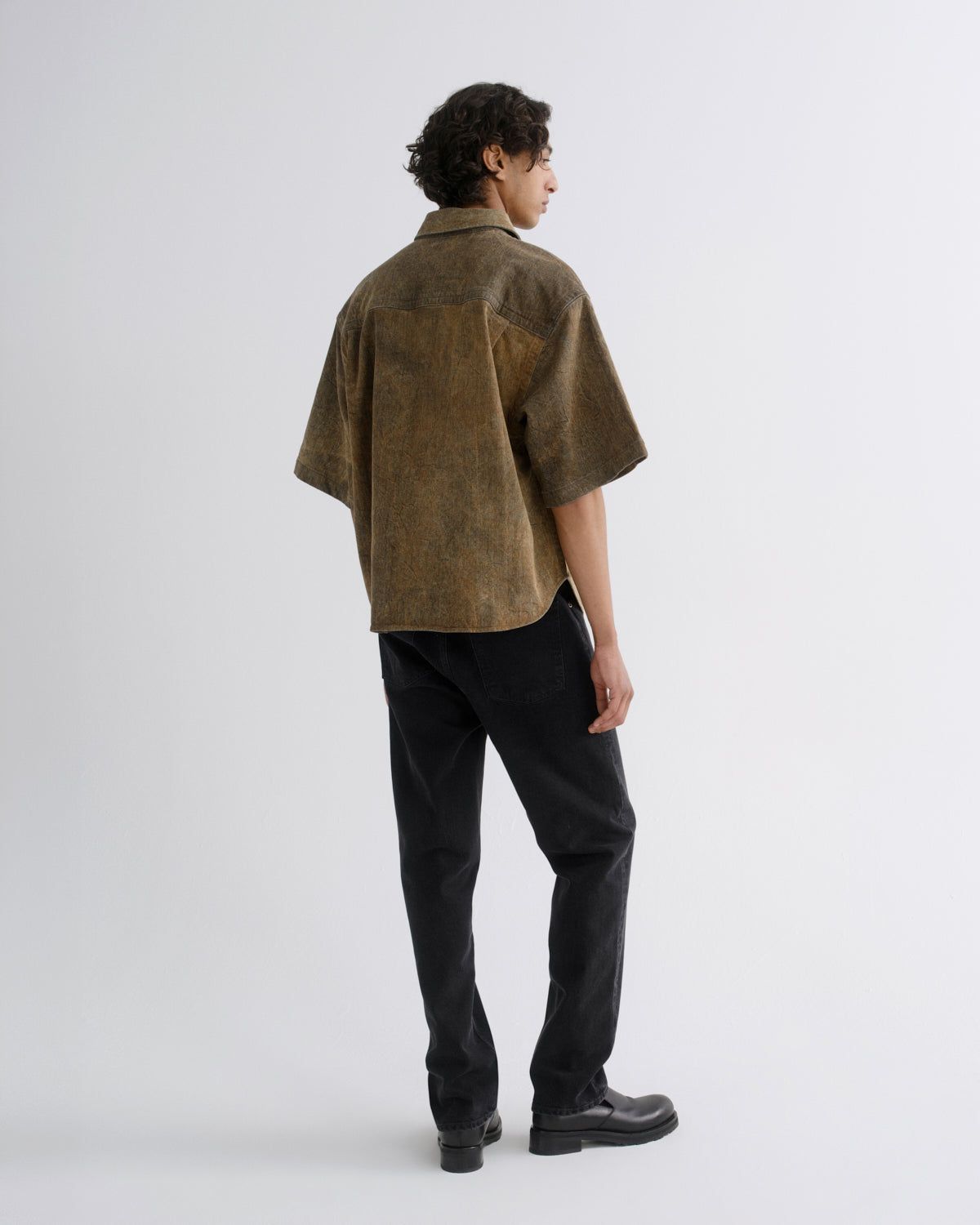 A man is shown from the back wearing a short-sleeved, textured brown shirt and black pants against a plain light gray studio background.
