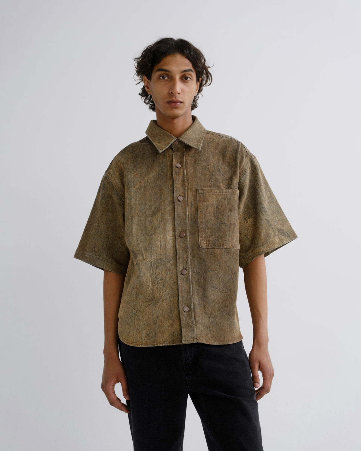 A model wearing a short-sleeved button-up shirt with a textured, mottled brown and beige pattern, paired with black pants, shot against a plain light gray background.