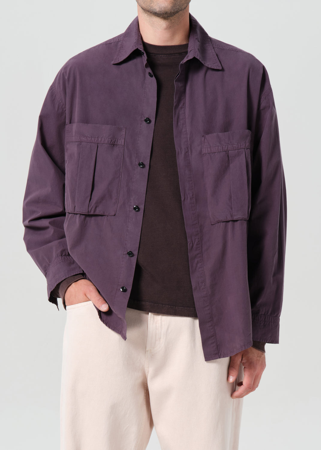 Person wearing a purple jacket over a dark shirt with beige pants on a light gray background