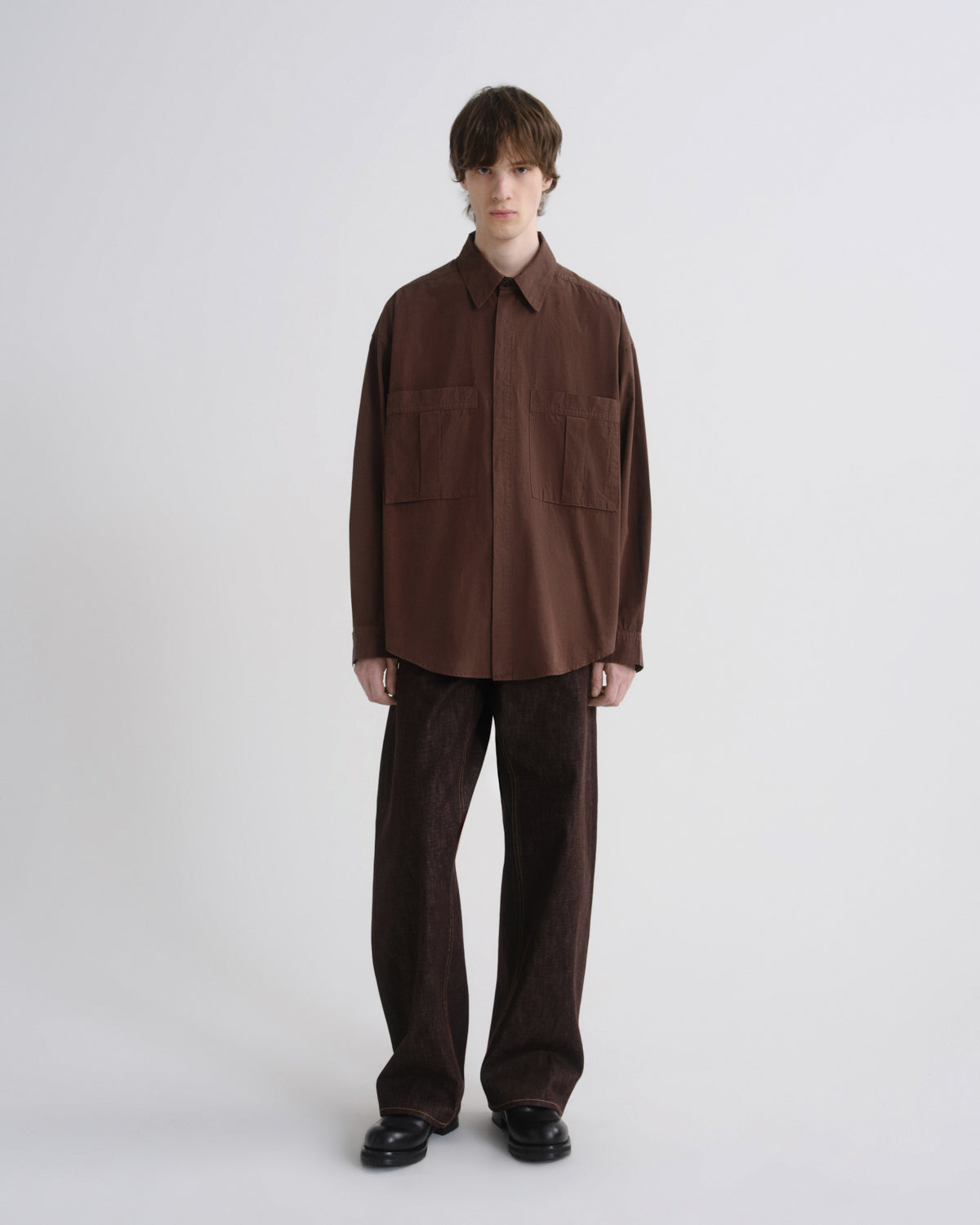 A model stands against a light gray studio background wearing a dark brown button-up shirt with two chest pockets and matching dark brown wide-leg trousers.