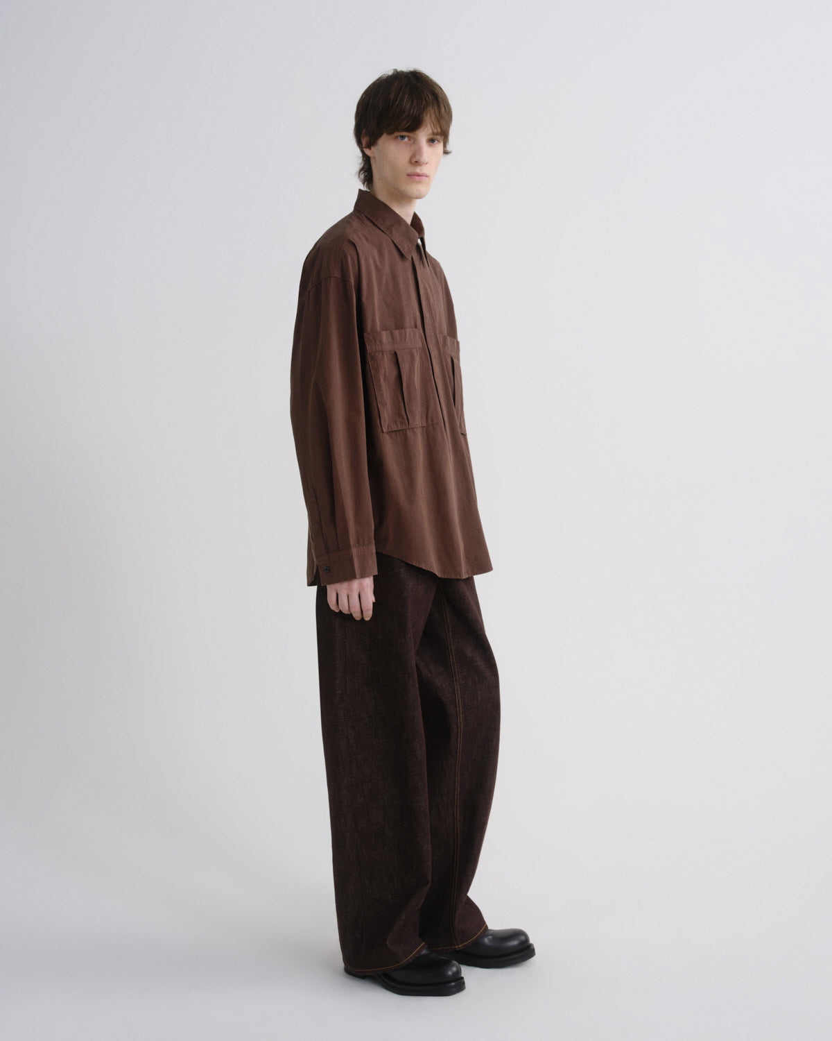A model stands in profile wearing a brown button-up shirt with chest pockets and matching dark brown wide-leg trousers against a plain light gray studio background.