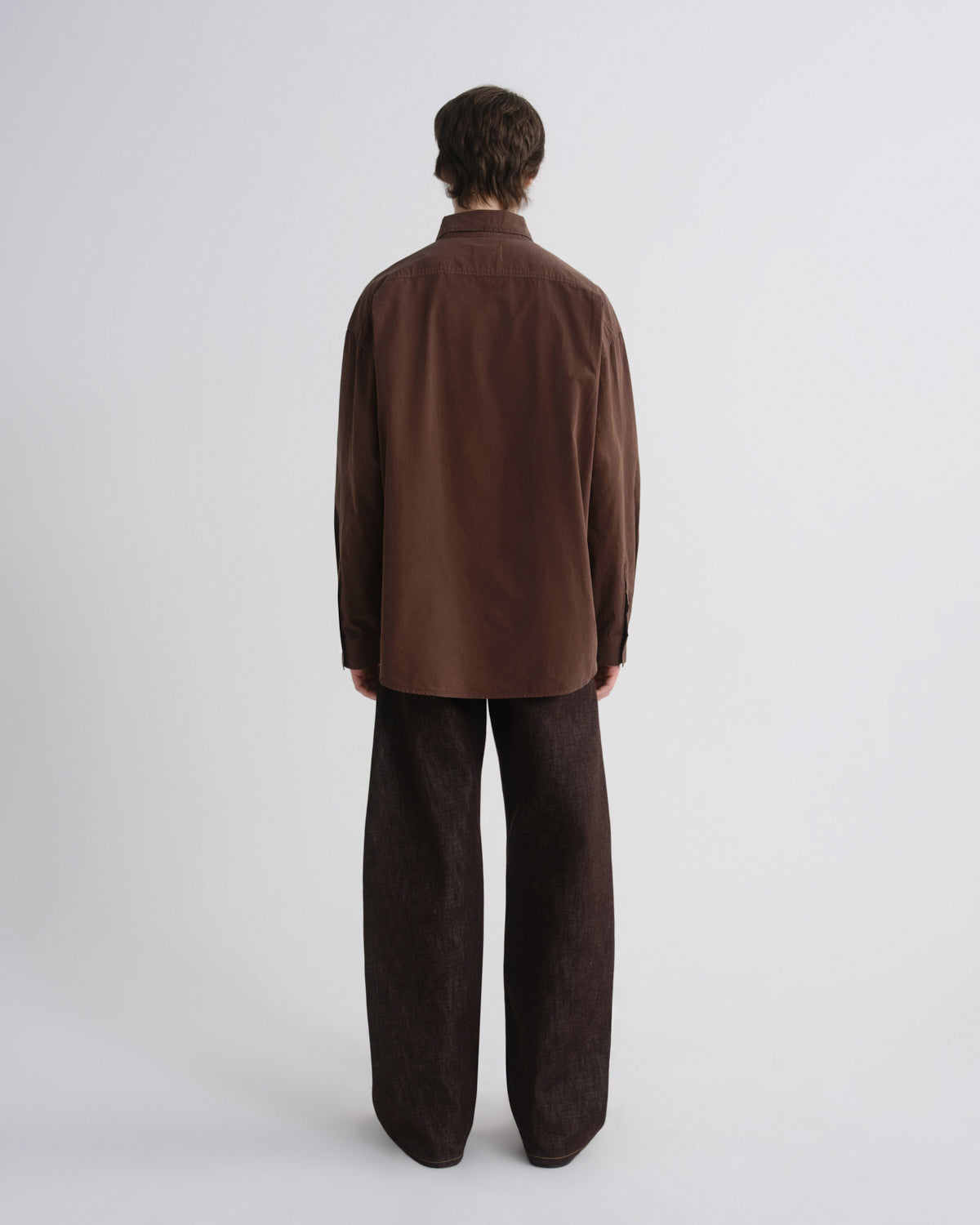 A model viewed from the back wearing a dark brown corduroy shirt and matching dark brown corduroy trousers, set against a plain light gray studio background.