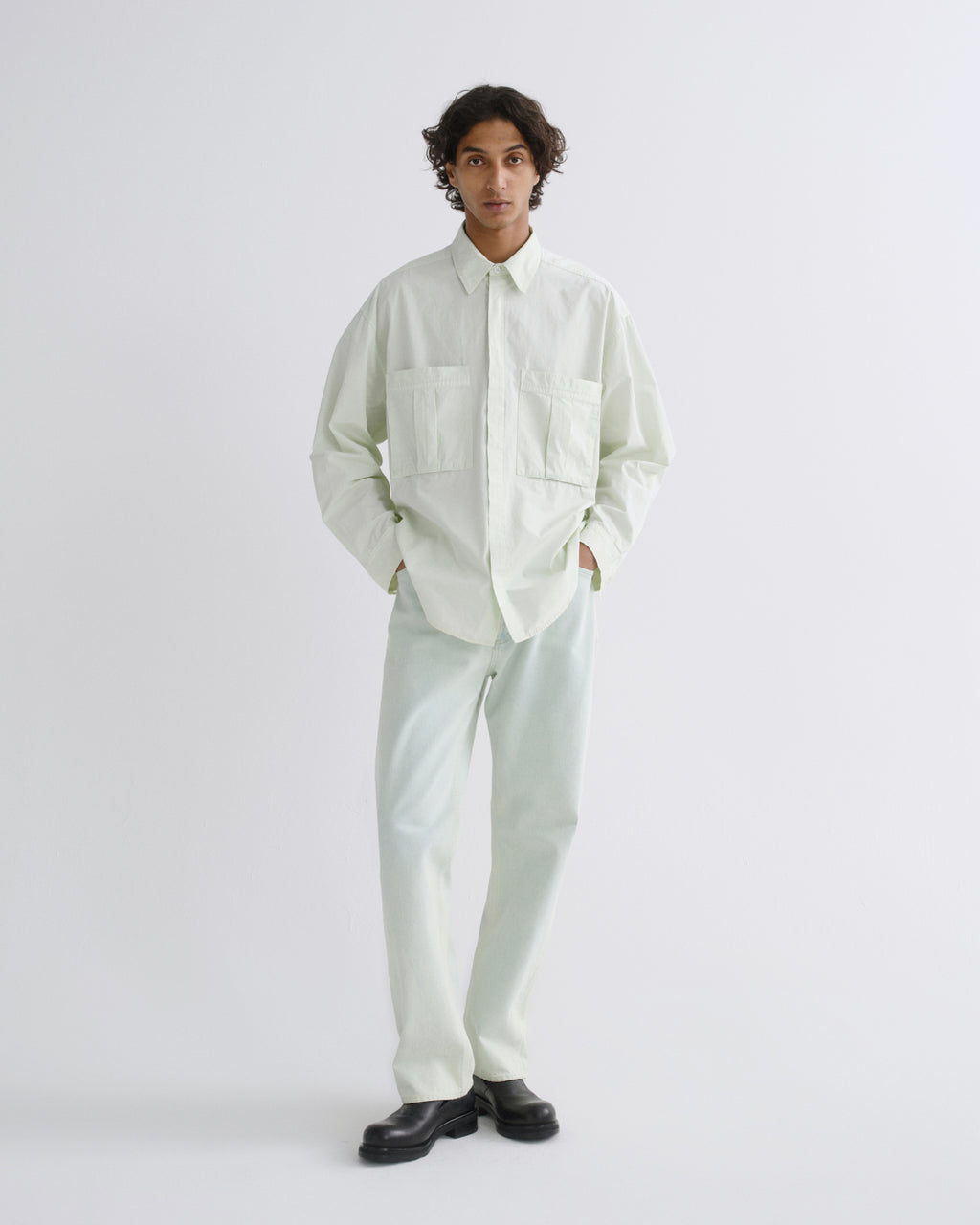 A model stands against a light gray studio background wearing a pale mint green long-sleeve button-up shirt with two chest pockets, matching light mint green trousers, and black leather shoes.