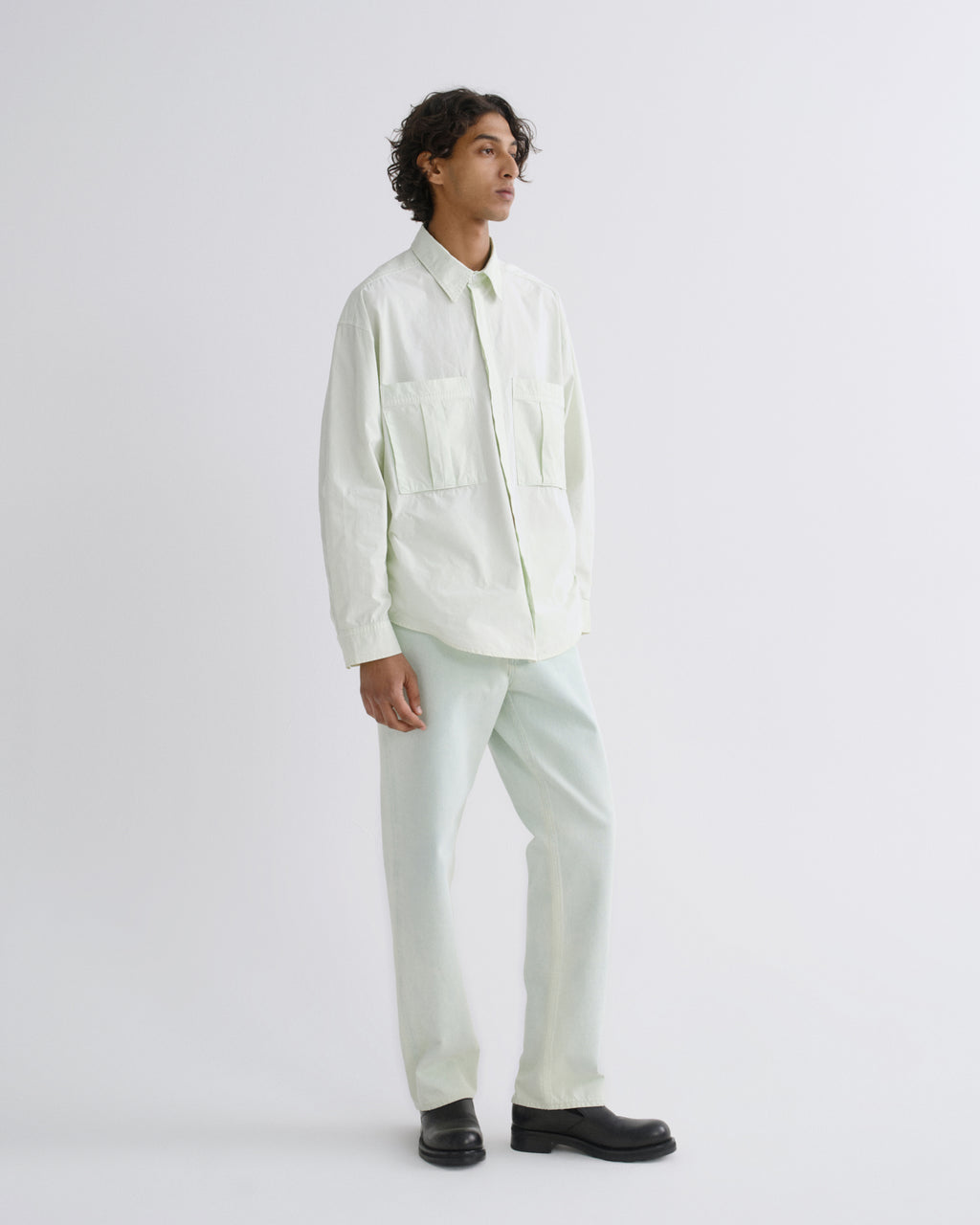 A man wearing a light cream long-sleeve button-up shirt with two chest pockets and matching light cream trousers, paired with black shoes, standing against a plain light gray studio background.