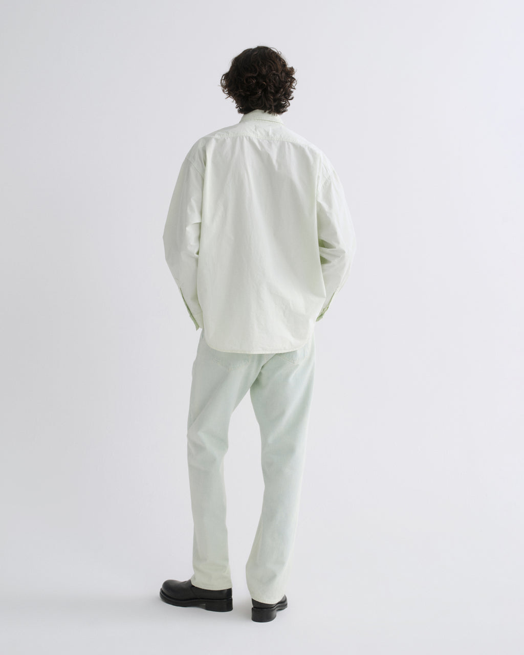 A man viewed from behind wearing a light sage green button-down shirt and matching light sage green trousers, paired with black shoes, against a plain white studio background.