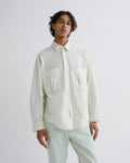 A model wearing a light sage green long-sleeve button-up shirt with two chest pockets and light sage green trousers, photographed against a neutral light gray studio background.