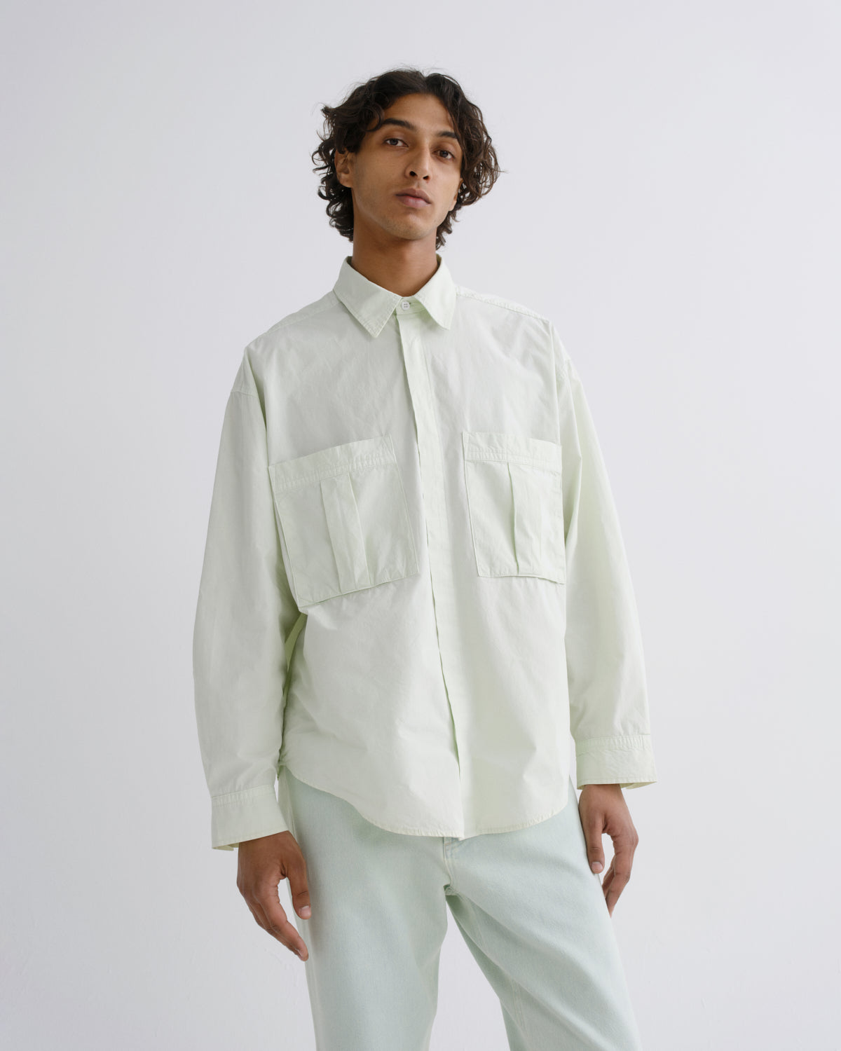 A model wearing a light sage green long-sleeve button-up shirt with two chest pockets and light sage green trousers, photographed against a neutral light gray studio background.