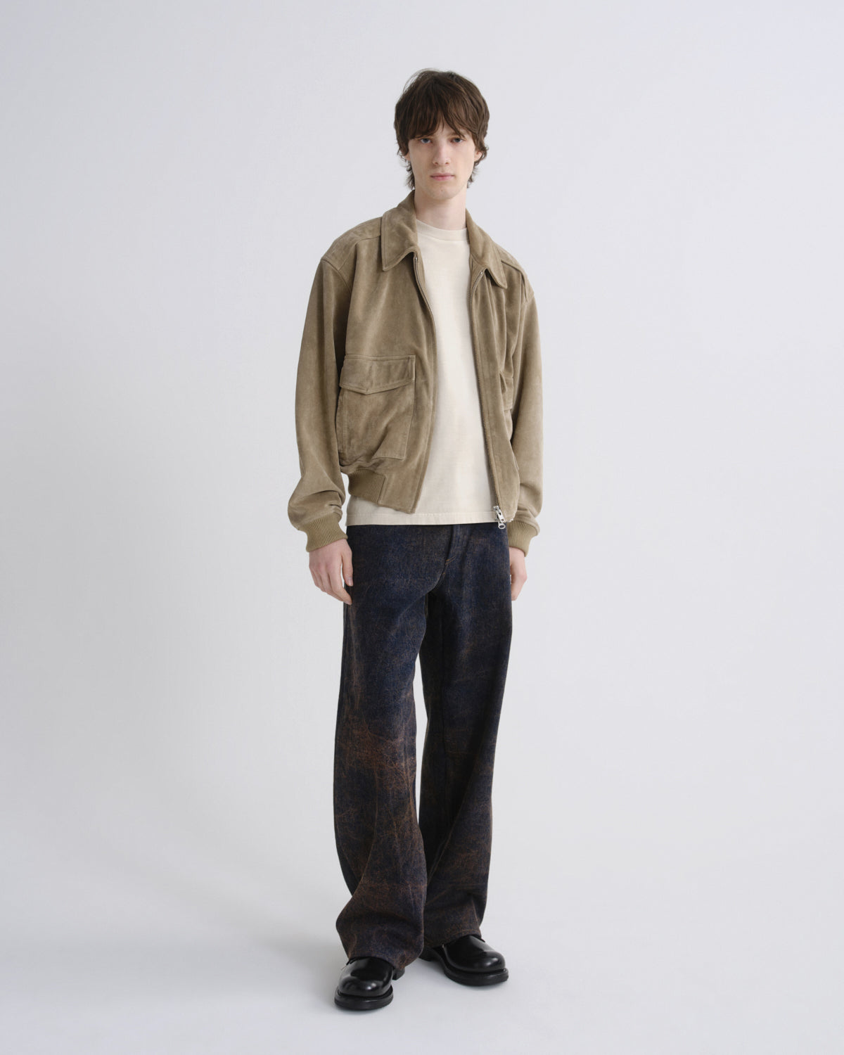 A model stands against a light gray studio background wearing a beige corduroy jacket over a cream turtleneck, dark blue patterned trousers, and black shoes.