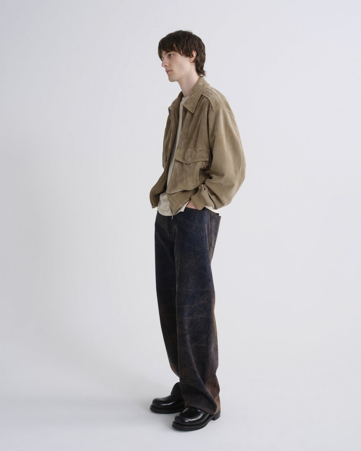 A model stands in profile wearing a light brown corduroy jacket over a white shirt, paired with dark indigo corduroy trousers and black leather loafers, against a plain light gray studio background.