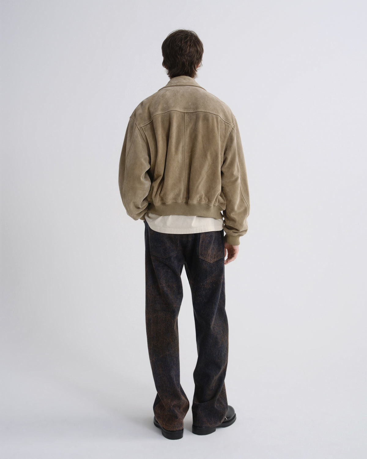 A model viewed from the back wearing a light brown suede bomber jacket over a cream shirt and dark washed denim jeans, shot against a plain light gray studio background.