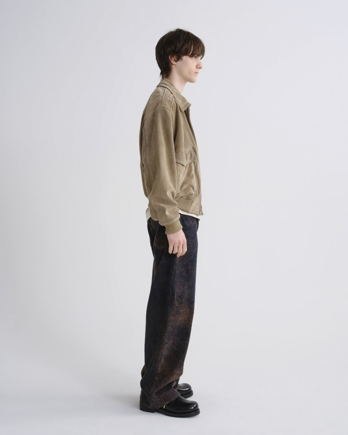 A model shown in profile wearing a light brown corduroy jacket, dark washed jeans, and black leather shoes against a plain light gray studio background.