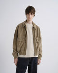 A model wearing a light brown suede utility jacket over a cream crewneck sweater and dark blue jeans, standing against a light gray studio background.