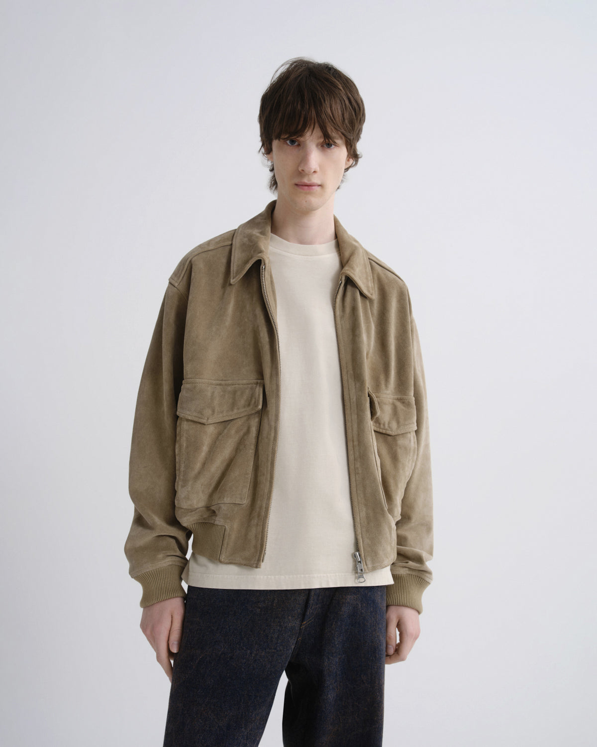 A model wearing a light brown suede utility jacket over a cream crewneck sweater and dark blue jeans, standing against a light gray studio background.