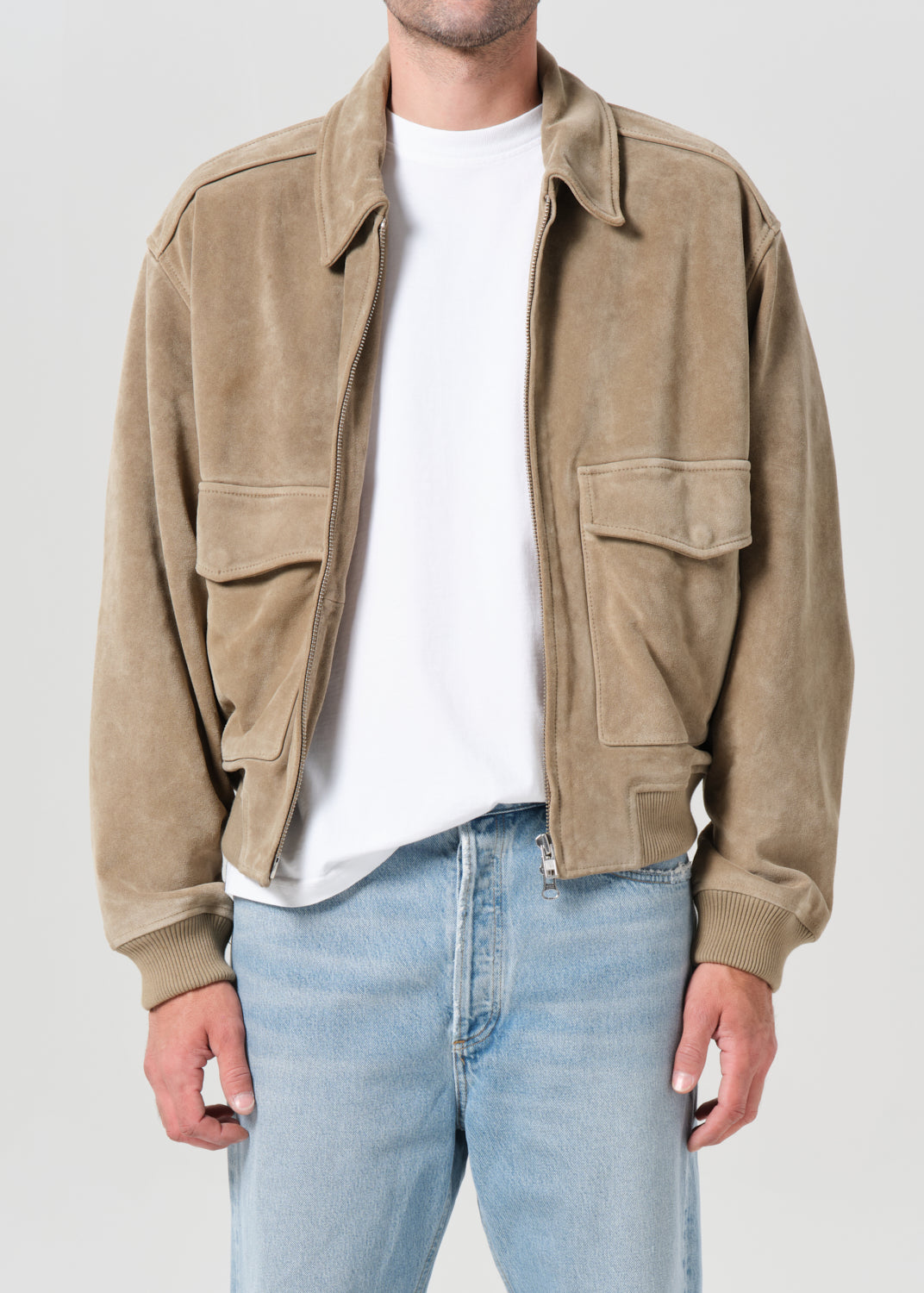 Person wearing a beige jacket, white t-shirt, and light blue jeans on a plain background