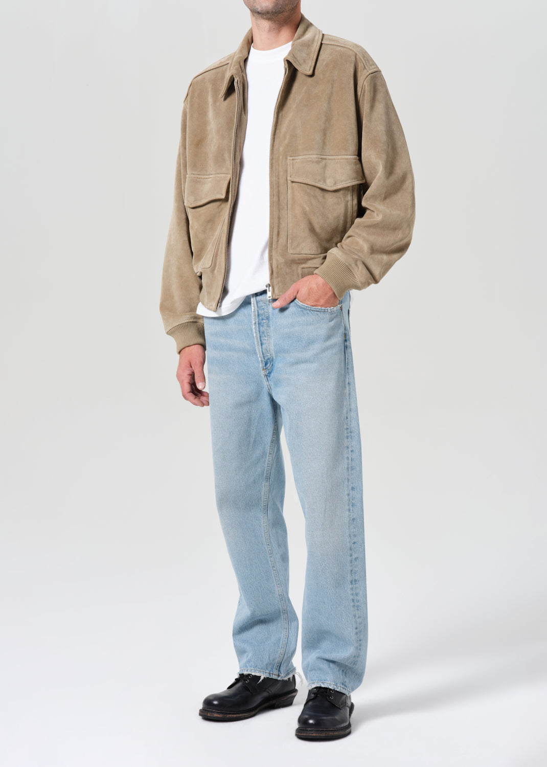 Person wearing a beige jacket, white shirt, light blue jeans, and black shoes on a white background