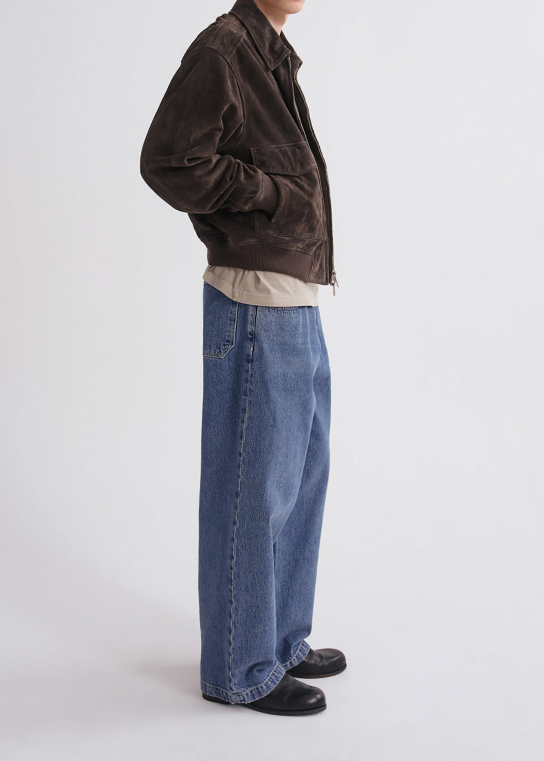 A side-profile studio shot of a person wearing a dark brown corduroy jacket, a beige t-shirt, wide-leg blue jeans, and black shoes against a grey background.