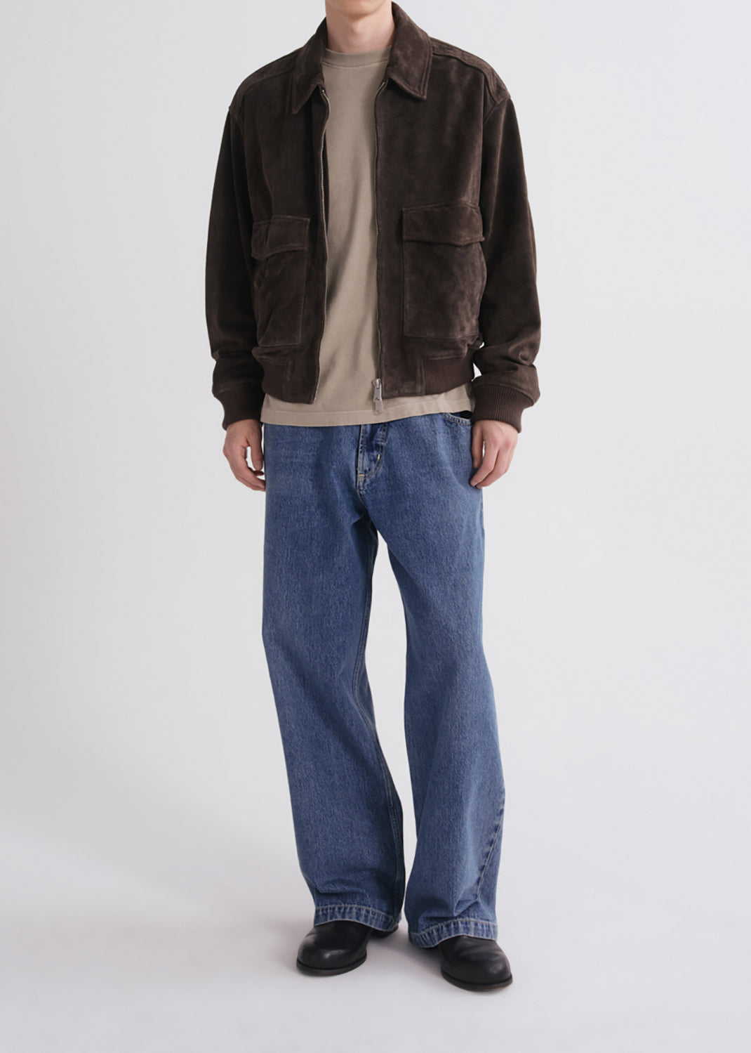 A model wearing a dark brown corduroy jacket over a beige t-shirt, paired with wide-leg blue jeans and black shoes, standing against a grey background.