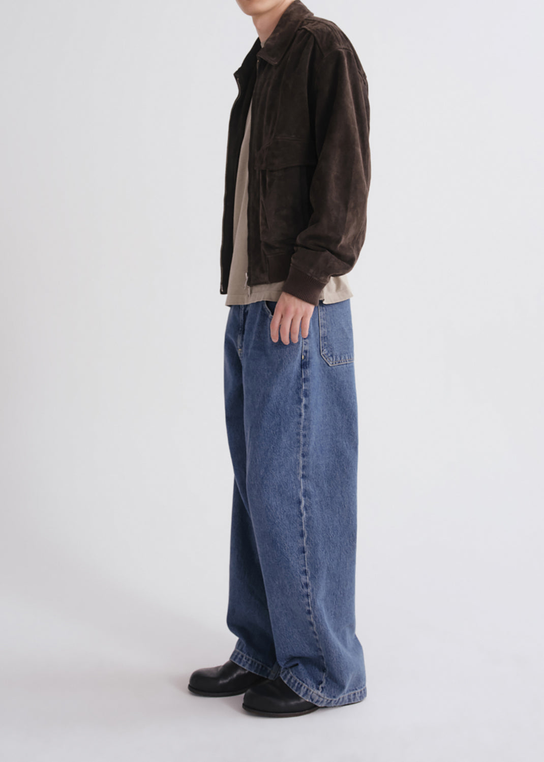 A model shown from the neck down wearing a dark brown corduroy jacket, a beige t-shirt, wide-leg blue denim jeans, and black leather shoes, standing against a grey background.