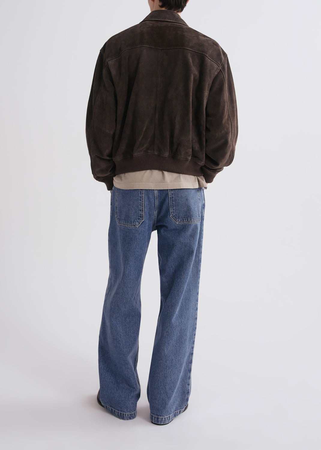 A person shown from the back wearing a dark brown suede jacket, a beige shirt, and wide-leg blue jeans against a grey background.