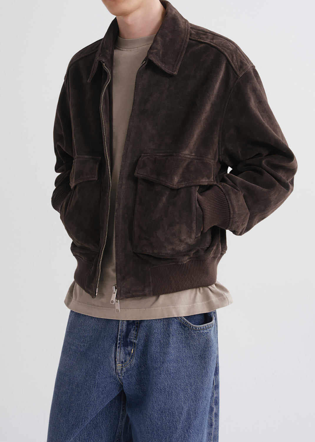 A person wearing a dark brown suede jacket over a beige t-shirt and blue jeans, shown from the neck down against a grey background.