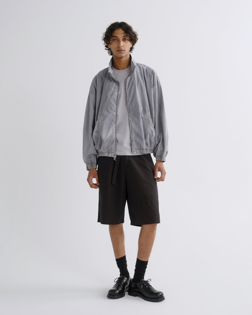 A model stands against a light gray studio background wearing a light gray zip-up jacket over a white t-shirt, black shorts, black socks, and black leather shoes.
