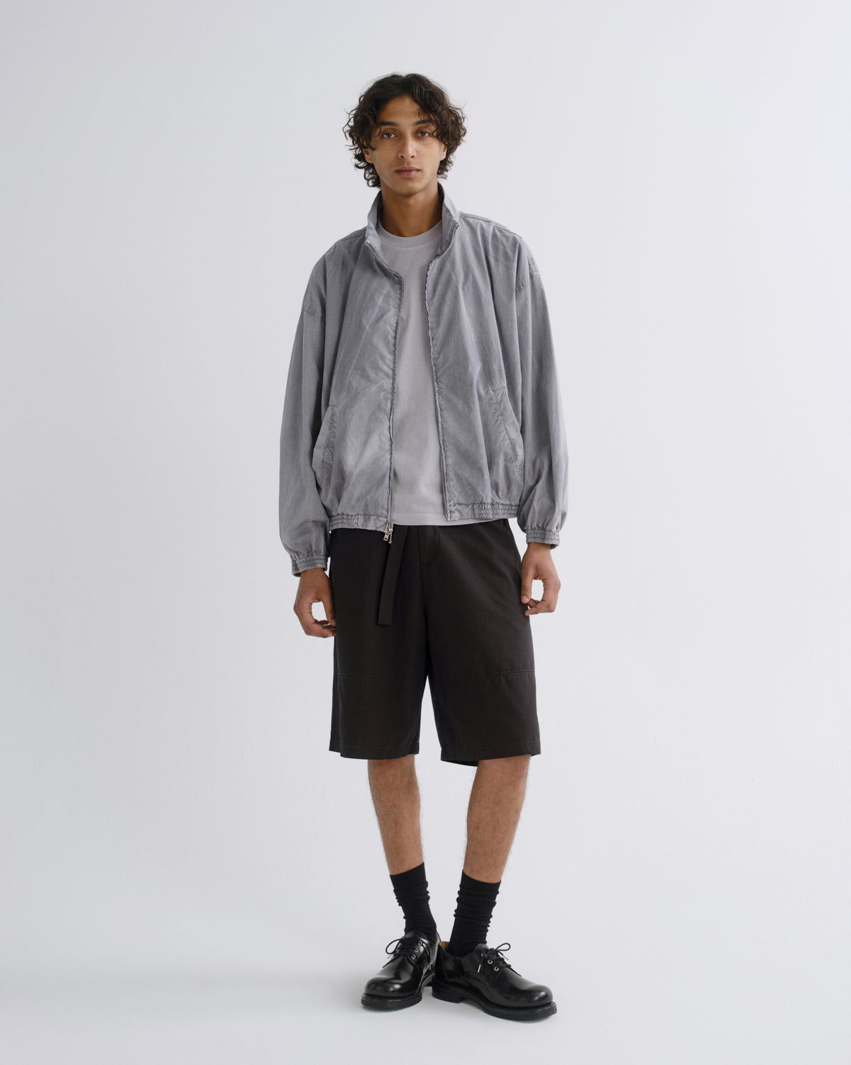A model stands against a light gray studio background wearing a light gray zip-up jacket over a white t-shirt, black shorts, black socks, and black leather shoes.