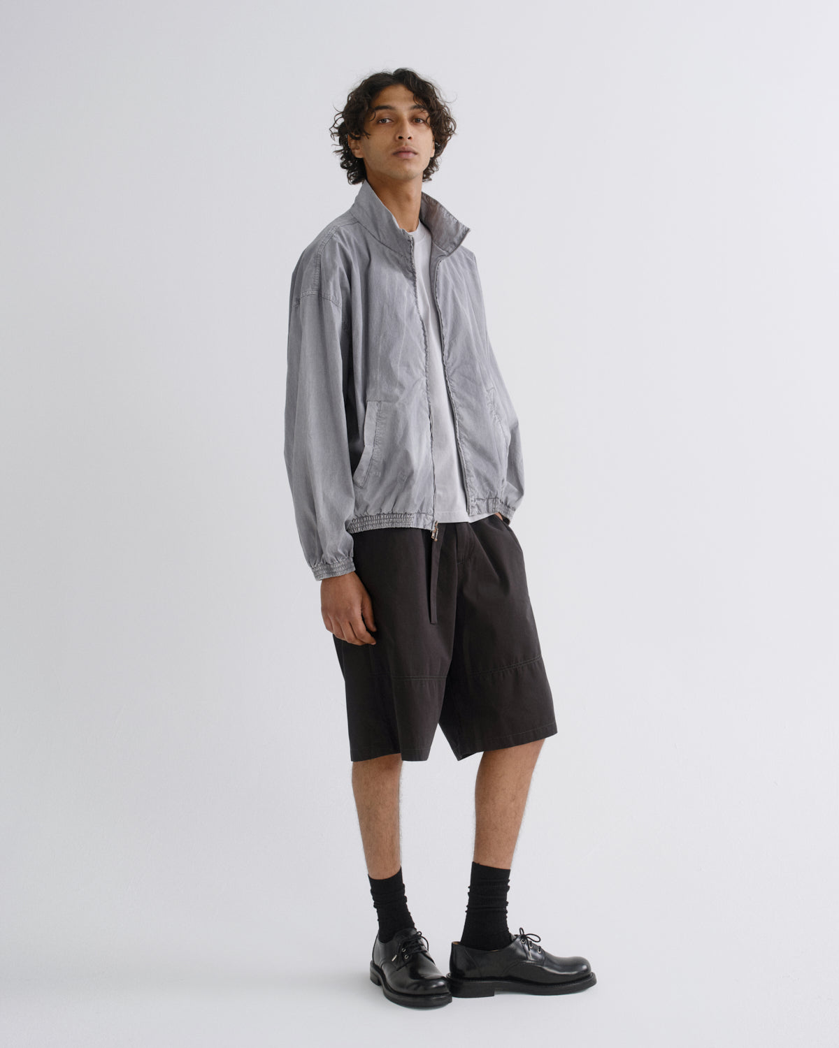 A model stands in a studio wearing a light gray lightweight zip-up jacket over a white shirt, paired with black drawstring shorts, black socks, and black leather shoes against a light gray background.