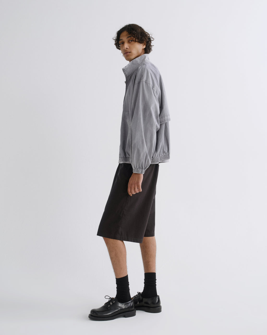 A model stands in profile wearing a light gray zip-up jacket, black knee-length shorts, black socks, and black leather shoes against a plain light gray studio background.