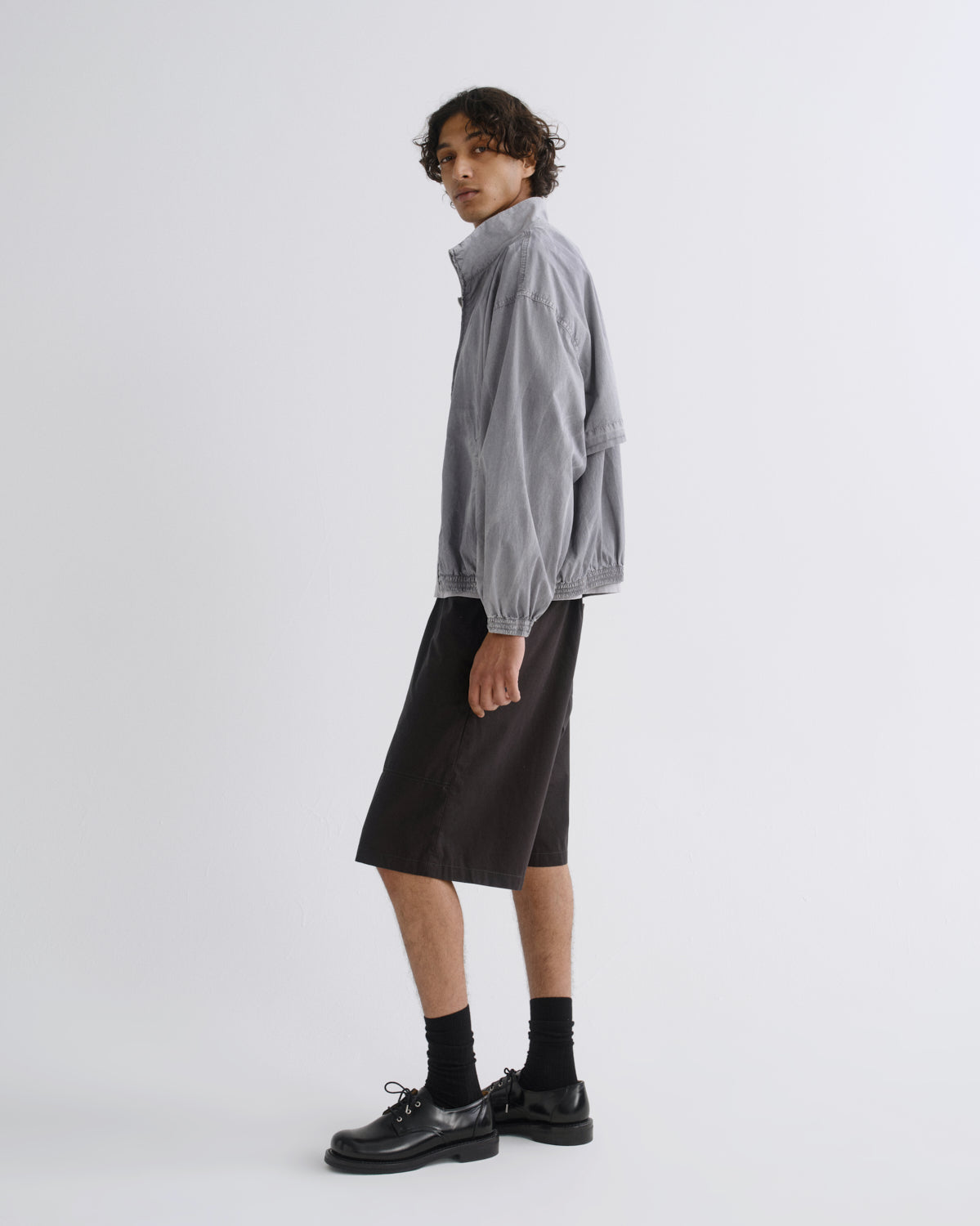A model stands in profile wearing a light gray zip-up jacket, black knee-length shorts, black socks, and black leather shoes against a plain light gray studio background.
