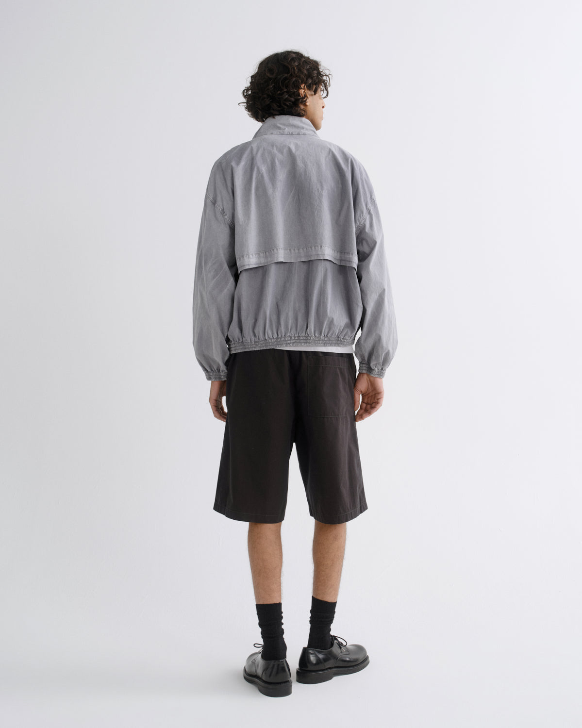 A model shown from the back wearing a light gray two-piece jacket and dark brown shorts, standing against a plain light gray studio background.