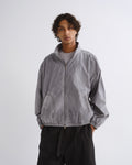 A man wearing a light gray zip-up windbreaker jacket over a gray t-shirt and black pants, standing against a light gray studio background.