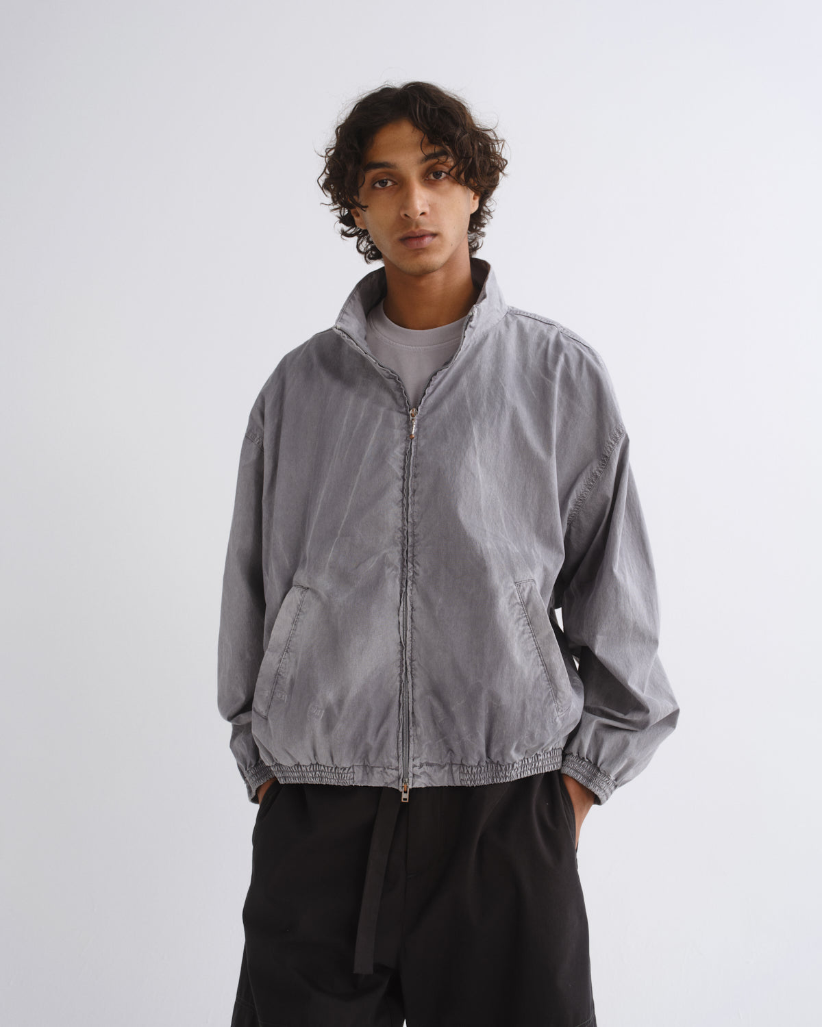 A man wearing a light gray zip-up windbreaker jacket over a gray t-shirt and black pants, standing against a light gray studio background.