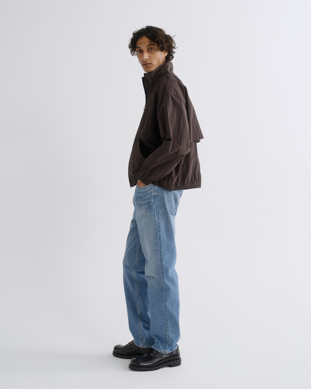 A model wearing a dark brown lightweight jacket, light blue wide-leg jeans, and black leather shoes, standing in profile against a light gray studio background.