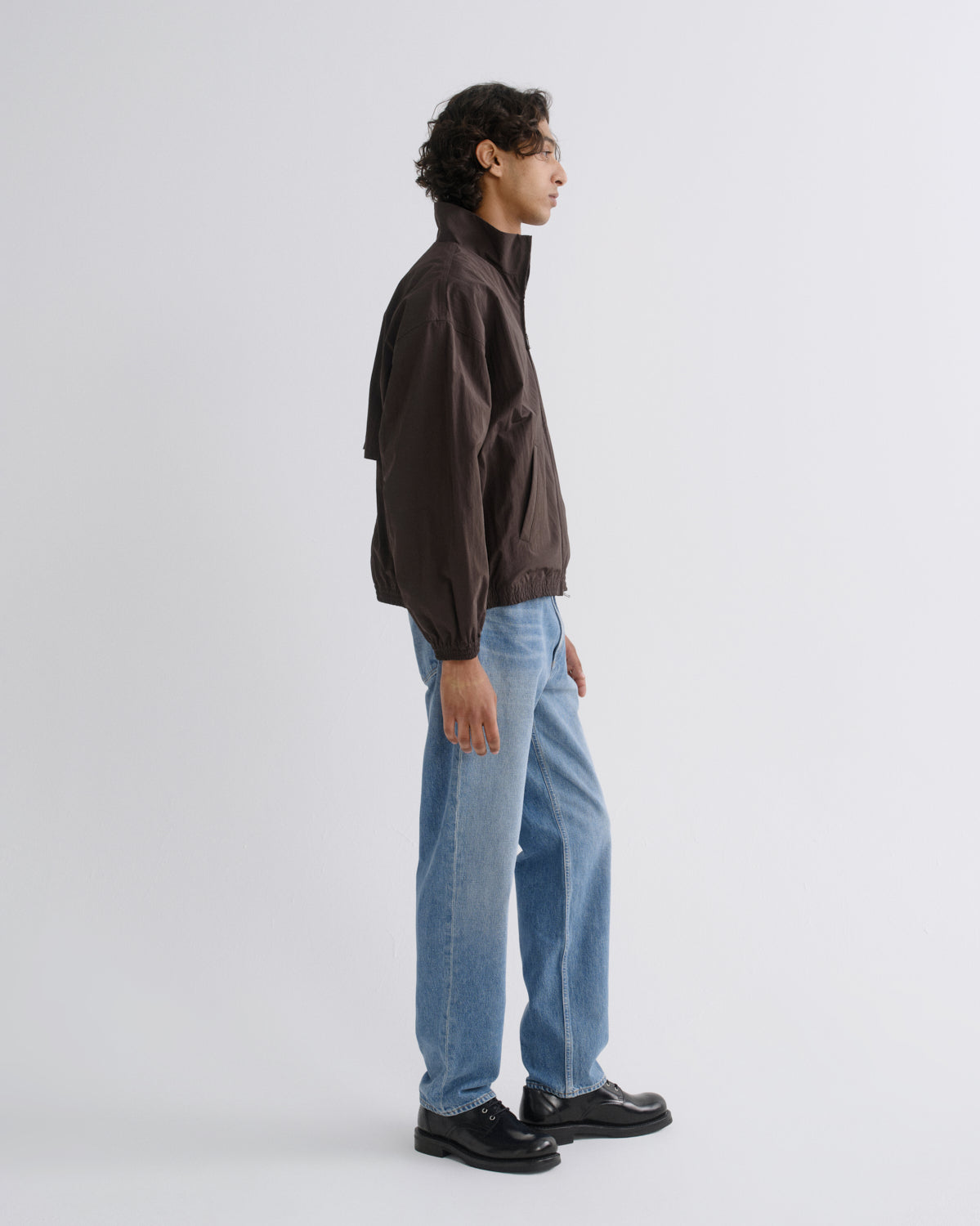 A man in profile wearing a dark brown lightweight jacket, light blue jeans, and black leather shoes, standing against a plain light gray studio background.