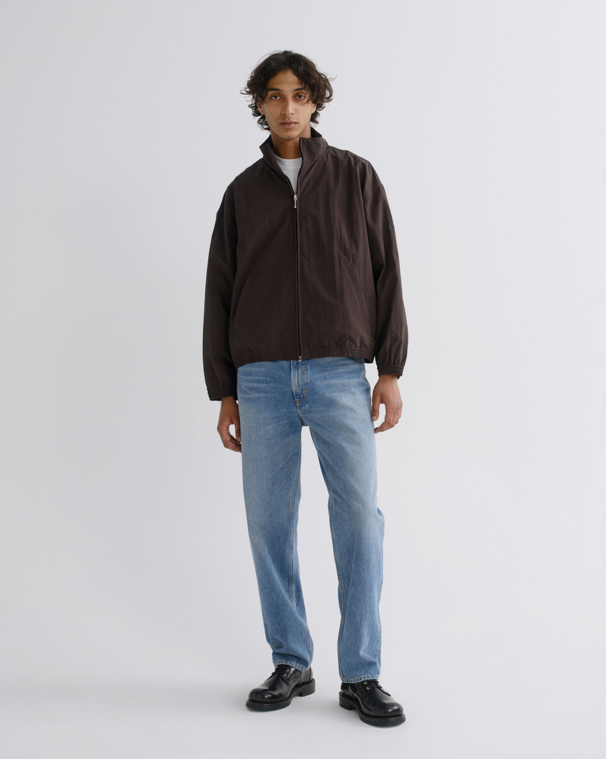 A model stands against a light gray studio background wearing a dark brown zip-up jacket, light blue jeans, and black shoes.