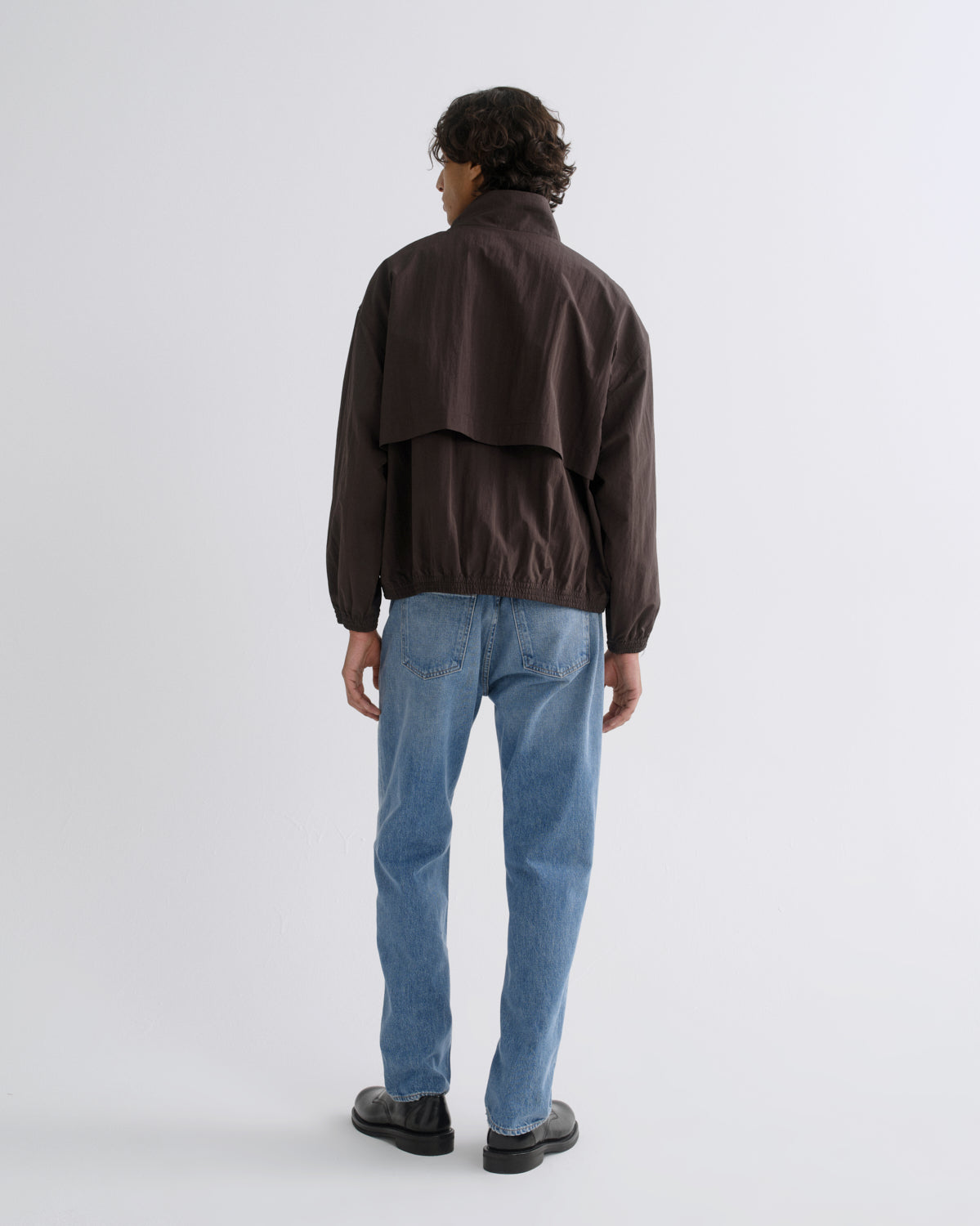A man viewed from behind wearing a dark brown jacket, light blue jeans, and black shoes, standing against a light gray studio background.