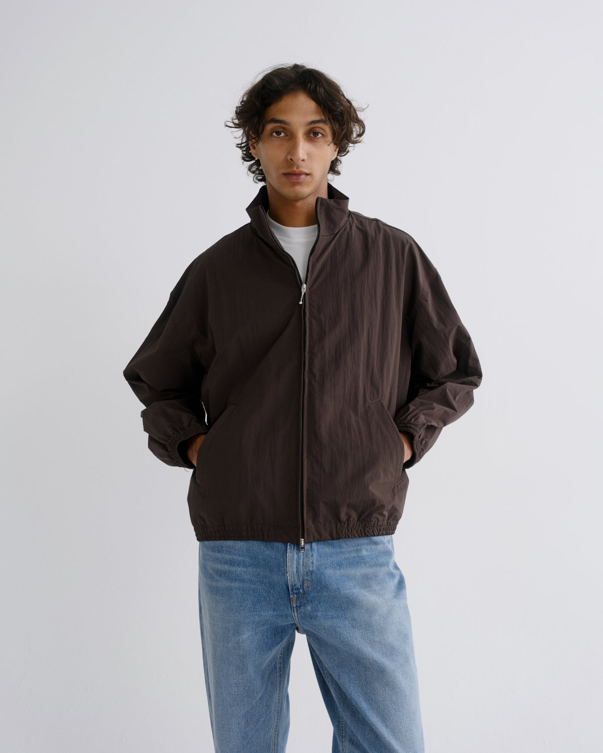 A man wearing a dark brown lightweight zip-up jacket over a white t-shirt and light blue jeans, standing against a plain light gray studio background.
