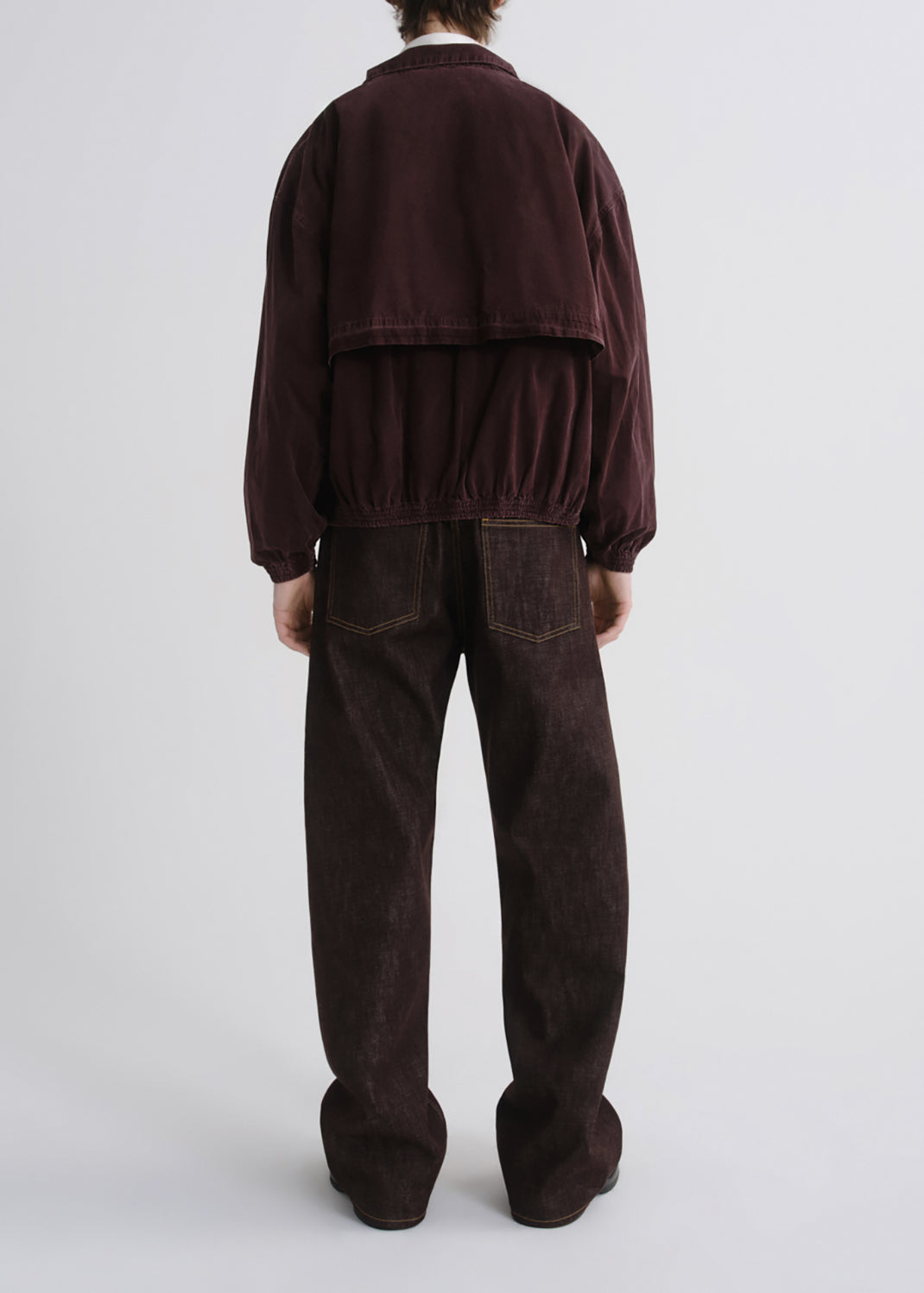 A model viewed from the back wearing a dark burgundy corduroy jacket with a cropped, layered back design and dark brown corduroy pants, set against a grey background.