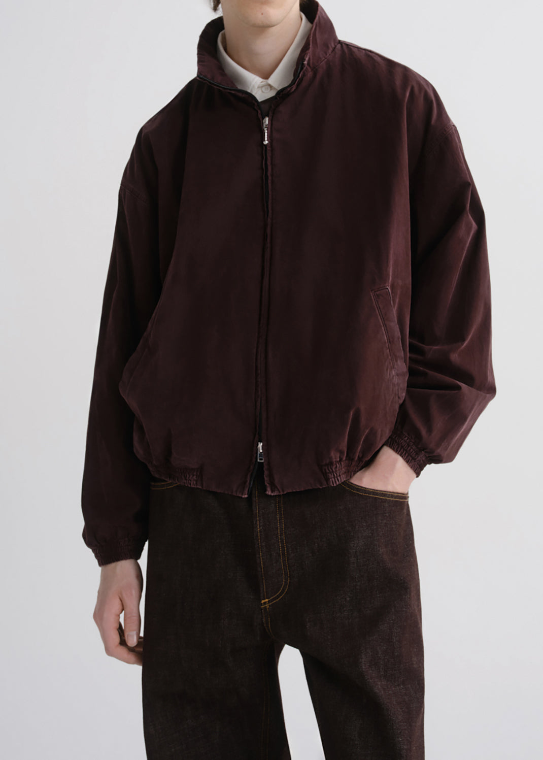 A person wearing a dark burgundy zip-up jacket over a light collared shirt and dark brown pants, photographed against a grey background.