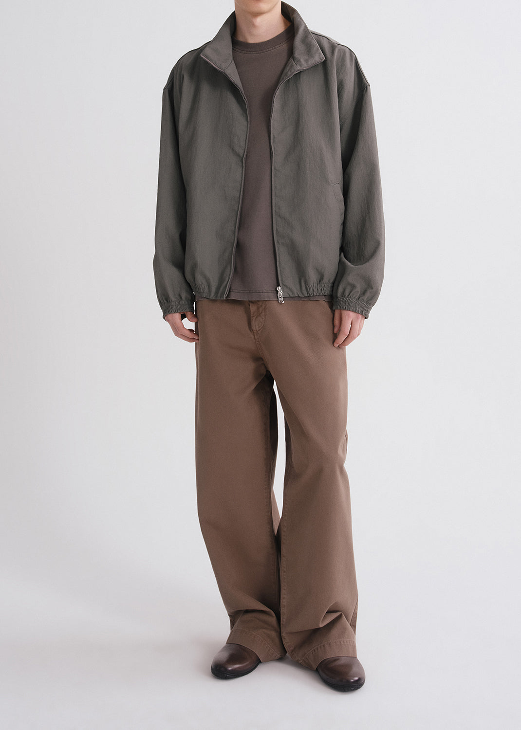 Person wearing green jacket over brown shirt and brown pants on a grey background