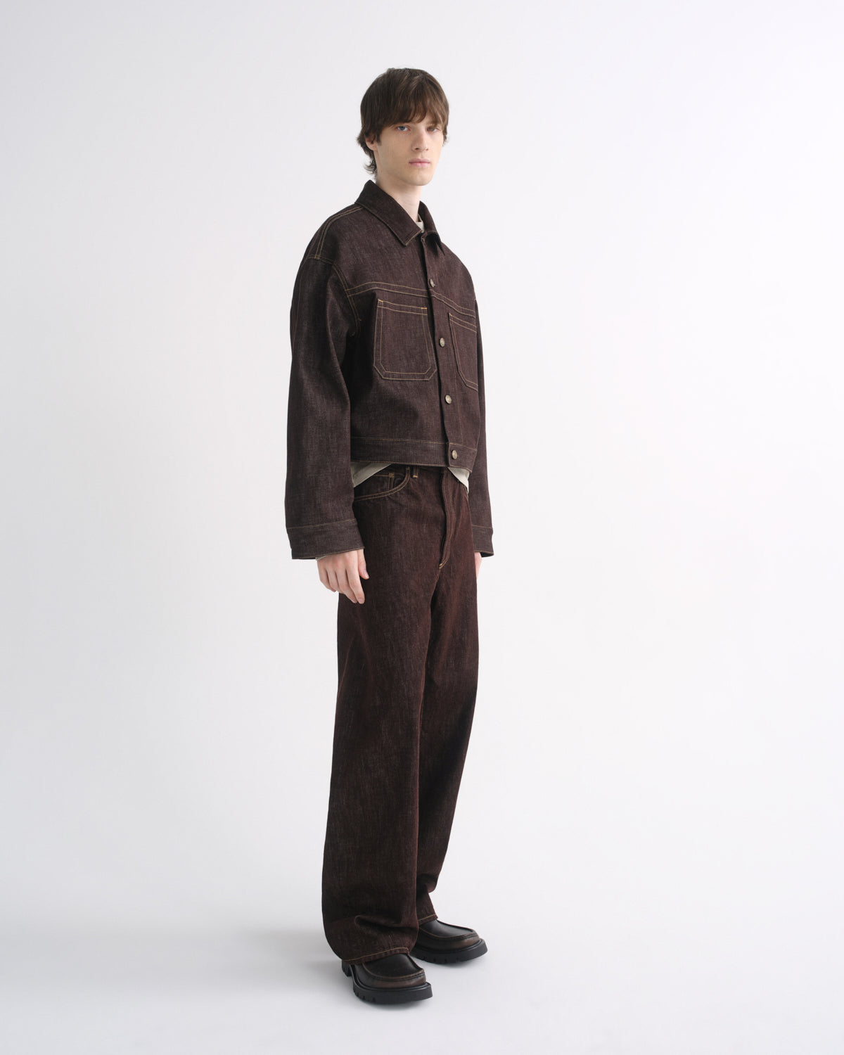 Person wearing a dark brown denim jacket with matching dark brown pants and black shoes, side view, on a grey background