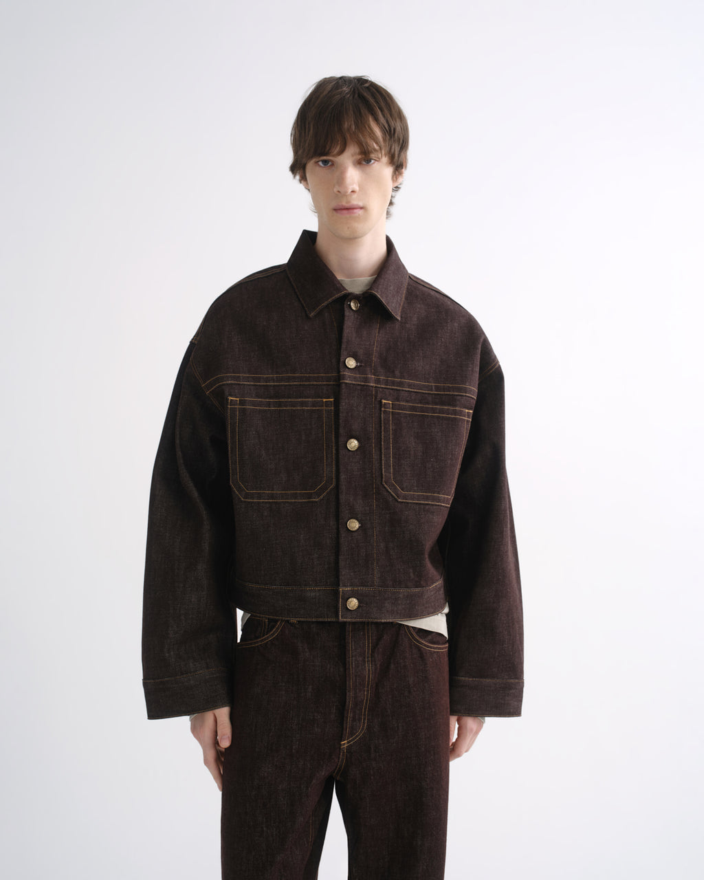 Person wearing a dark brown denim jacket with matching dark brown pants and black shoes on a grey background