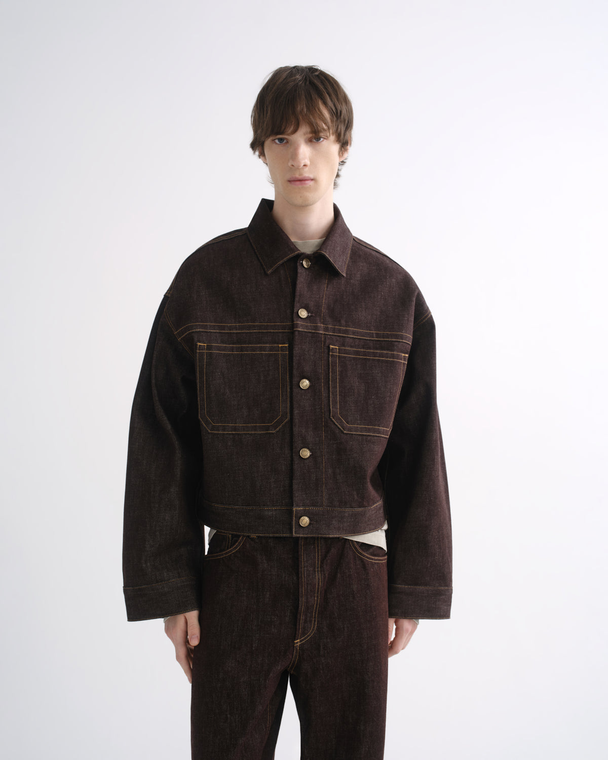 Person wearing a dark brown denim jacket with matching dark brown pants and black shoes on a grey background